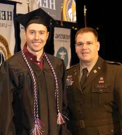 Herzing University graduate in cap and gown posing with a military service member in uniform during a graduation ceremony.